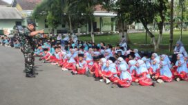 Foto: Tampak seorang prajurit TNI berdiri di halaman sebuah markas dan berinteraksi dengan sekelompok besar siswa sekolah dasar/madrasah yang duduk berbaris di atas aspal. Anak-anak mengenakan seragam bernuansa biru muda dan merah