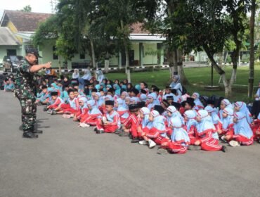 Foto: Tampak seorang prajurit TNI berdiri di halaman sebuah markas dan berinteraksi dengan sekelompok besar siswa sekolah dasar/madrasah yang duduk berbaris di atas aspal. Anak-anak mengenakan seragam bernuansa biru muda dan merah
