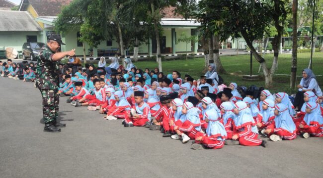 Foto: Tampak seorang prajurit TNI berdiri di halaman sebuah markas dan berinteraksi dengan sekelompok besar siswa sekolah dasar/madrasah yang duduk berbaris di atas aspal. Anak-anak mengenakan seragam bernuansa biru muda dan merah
