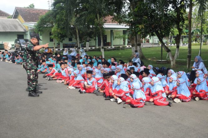 
					Foto: Tampak seorang prajurit TNI berdiri di halaman sebuah markas dan berinteraksi dengan sekelompok besar siswa sekolah dasar/madrasah yang duduk berbaris di atas aspal. Anak-anak mengenakan seragam bernuansa biru muda dan merah